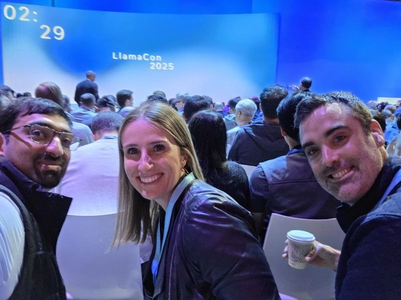 Three attendees taking a selfie with the LlamaCon stage and audience behind them.